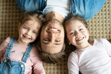 Top view of handsome young father and his cute little daughters looking at camera and smiling, lying on floor.