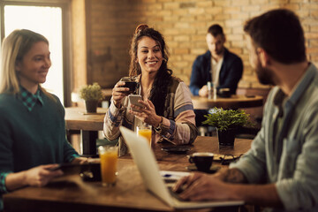 Happy woman using smart phone and drinking coffee while being with her friends in a cafe.