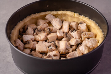 On the kitchen table there is a mold with shortcrust pastry, inside are mushrooms and chicken pieces.