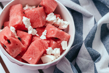 Delicious summer salad with slices of watermelon and feta cheese on white bowl with a striped towel on a wooden white background