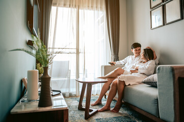 Asian couple watching television sitting on a sofa in the living room at home. Man is  holding a...
