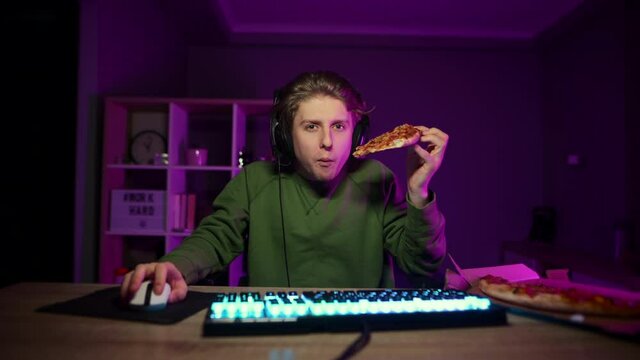 Focused young man in a headset plays video games at home on the computer at night and eats a slice of pizza, looking focused at the screen.