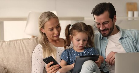 Addicted with modern gadgets family sit on sofa spend time together using diverse device. Small kid play videogame on tablet rest at home with parents holding phone and pc. Modern tech overuse concept - Powered by Adobe