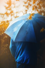 A man in a blue jacket and with a blue umbrella in his hand walks alone in the park on an autumn sunny rainy day. October.