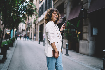 Cheerful hipster girl in electronic headphones enjoying music playlist from smartphone application, happy Caucaisan tourist received funny audio message using 4g internet on cellphone technology
