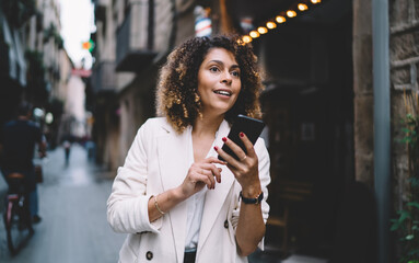 Millennial curly hipster girl with digital mobile phone in hands looking for something during travel sightseeing in city, charming female tourist using smartphone technology during solo journey