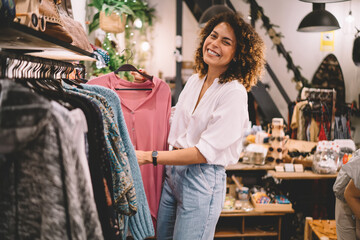 Portrait of joyful female customer enjoying weekend shopping in brand showroom, cheerful woman in trendy outfit buying stylish clothing and smiling at camera while posing near hangers with wear