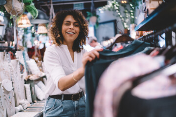 Fototapeta na wymiar Portrait of joyful female customer enjoying weekend shopping in brand showroom, cheerful woman in trendy outfit buying stylish clothing and smiling at camera while posing near hangers with wear