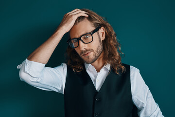 Romantic young man in elegant clothing and eyewear looking at camera and keeping hand in hair while...