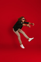 Full length of surprised young man in casual clothing making a face and keeping arms outstretched while hovering against red background - obrazy, fototapety, plakaty