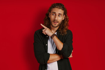 Good looking young man in casual clothing looking away and pointing copy space while standing against red background