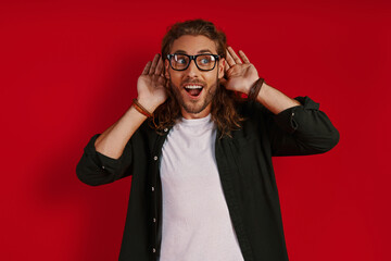 Curious young man in casual clothing keeping hands near ears and smiling while standing against red...