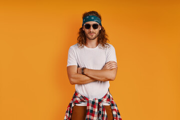 Stylish young man looking at camera and keeping arms crossed while standing against yellow background