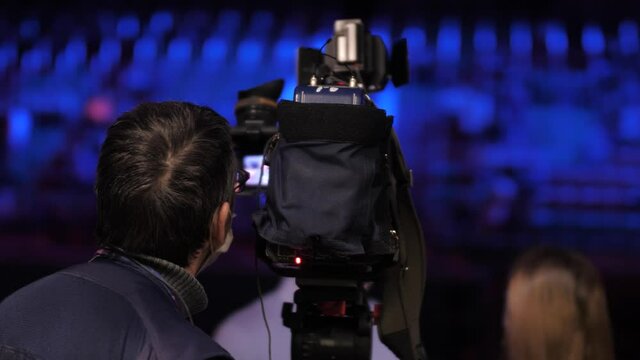 a cameraman filming an interview. Shooting from the back. In the background of the interview, a large stadium with rows of chairs