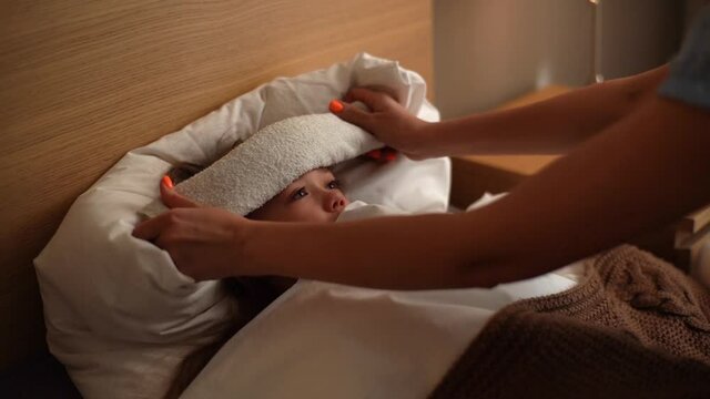 Young mother putting compress on forehead to bring down temperature of sick daughter with fever lying under blanket at home. Parent caring for sick child lying in bed with flu. Shooting in slow motion