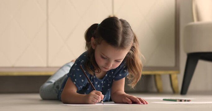 Little 4s girl with ponytails lying on warm floor in cozy comfort living room absorbed in favourite pastime holding pencil draw in sketchbook, create paintings. Hobby at home, creative kid concept