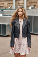 Young stylish woman in pink dress, black leather jacket and boots walking at urban location. Female model with long wavy blonde hair outdoor fashion look.