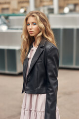 Fototapeta premium Young stylish teenager girl with blue eye arrows and long blonde wavy hair. Trendy fashionable female in pink dress, black leather jacket and high boots. Rock style woman outdoor portrait