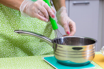 Gloved hands hold the whisk and stir the ingredients in the pan. The cooking process. Selective focus.