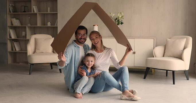Young happy spouses their little preschooler daughter sit on floor in modern light warm living room under carton roof smiling looking at camera. New dwelling symbol, portrait family homeowners concept