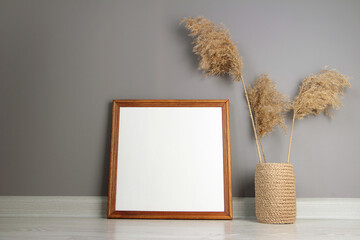 Empty mockup poster. Blank square picture in wooden frame  on background gray wall. Dried reed in a knitted jute vase. Minimal interior design. Modern style.