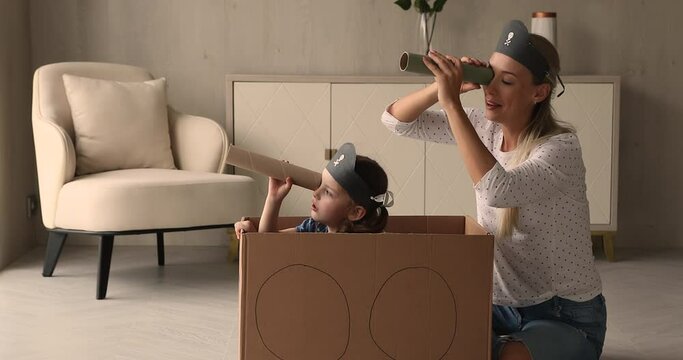 Little kid girl sit inside of carton box play pirates with mother, family holding paper tubes spyglasses watching into distance pretend be explorers. Funny playtime with children at home, fun concept
