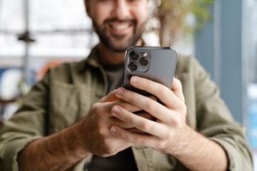 Unshaven happy man smiling and using mobile phone while sitting in cafe