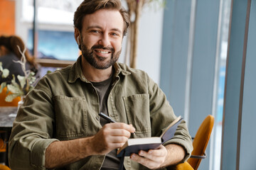 Unshaven happy man in earphones writing down notes in diary