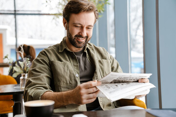 Unshaven smiling man in earphones reading newspaper in cafe