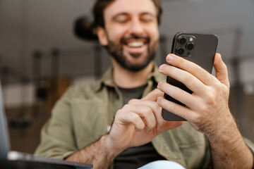 Unshaven happy man smiling and using mobile phone while sitting at cafe