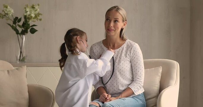 Adorable little wear white coat uniform play doctor take care of mother holds stethoscope listens to mom heartbeat. Funny playtime with kid at home, cardiologist profession in future, vocation concept