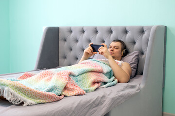 A young man in wireless headphones lies in a bed and watches video on his phone.