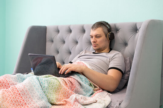 A Young Man Freelancer In Headphones Lies Reclining In A Bed Under A Multicolored Plaid And Watches Video Or Works On A Small Laptop.