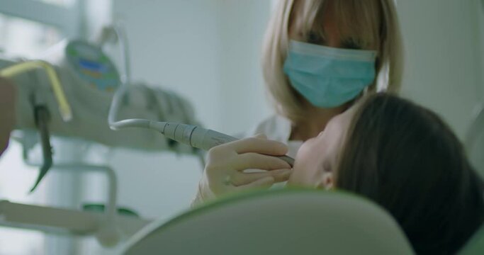 A young girl is sitting in the dentist chair and having her mouth open; the dentist is wearing a disposable mask and treating the girl’s teeth with a wire electrical tool.