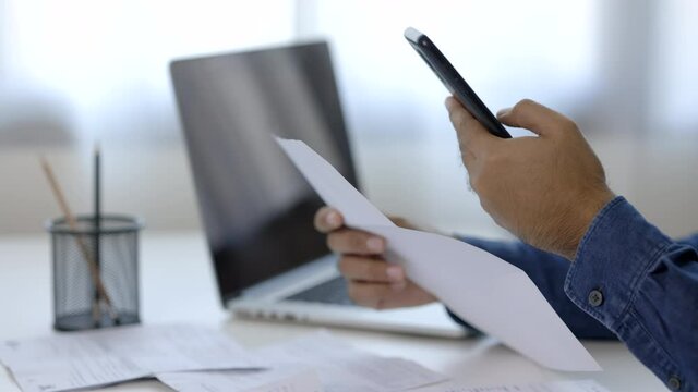 A young man is using a smartphone to scan the bill to calculate his income and expenses. In his hand there were bills to pay a lot in debt.
