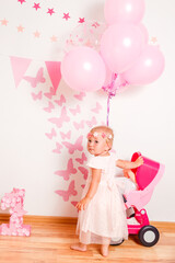 Happy blonde girl in a beautiful pink dress plays with doll's stroller at home. one-year photo shoot