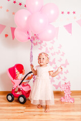 Happy blonde girl in a beautiful pink dress plays with doll's stroller at home. one-year photo shoot