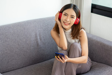 A young Asian woman happily listening to music on a sofa inside a home office using a wireless headphone, work from home concept.