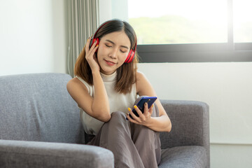 A young Asian woman happily listening to music on a sofa inside a home office using a wireless headphone, work from home concept.