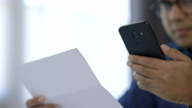 A young man is using a smartphone to scan the bill to calculate his income and expenses. In his hand there were bills to pay a lot in debt.
