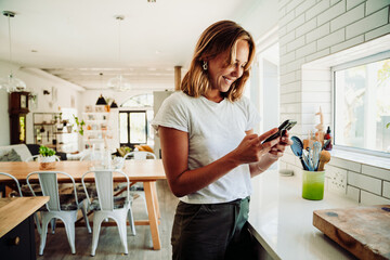 Caucasian female student working from home typing on cellular device standing in kitchen