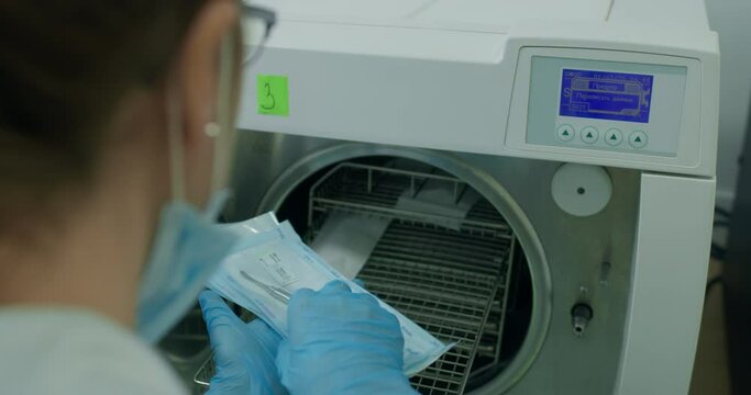 A medical worker is opening an autoclave machine with some metal containers, she is holding some plastic bags with the dental tools inside, she is putting the bags in the containers.