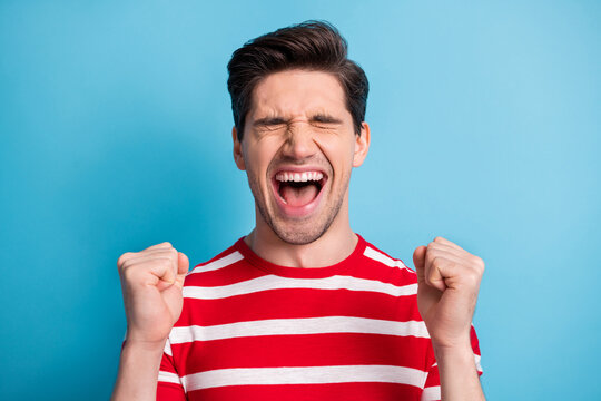 Photo of happy cheerful young man raise fists winner closed eyes scream luck isolated on blue color background