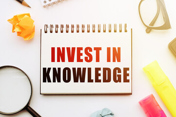 Text INVEST IN KNOWLEDGE in notebook on white table with office tools.