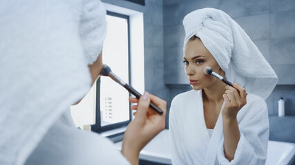 young woman applying face powder with cosmetic brush while looking at mirror in bathroom