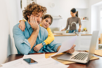 Young father stay at home trying to work on laptop while being distracted by his small daughter. Mother cooking in kitchen