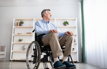 Low angle view of impaired male patient in wheelchair looking out window, feeling sad and desperate at home