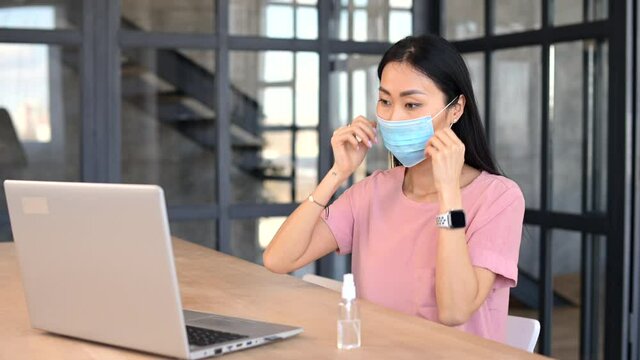 Asian female worker in medical mask sitting at the desk and typing on the laptop in office room protecting herself from getting virus, keeps positive, uses an antiseptic, works online, keeps distance