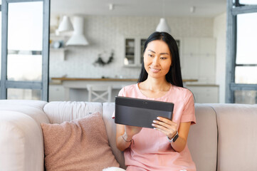 Portrait of young asian woman in a pink home T-shirt with black straight hair and snow-white smile, using modern grey digital tablet for texting, reading, watching video and surfing online at home - Powered by Adobe