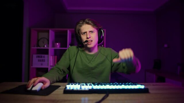 Upset male gamer playing video game on computer at home at night in room with purple light and showing thumbs down. Gamer shows a dislike